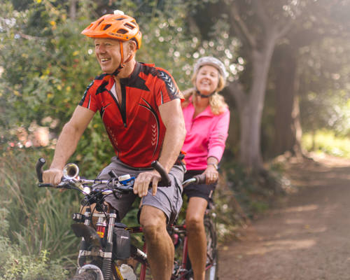 Smiling man and woman riding a tandem bike down a sunny path through the woods