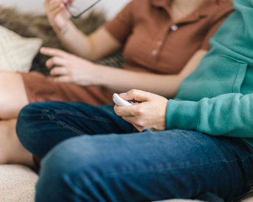 Two people sitting on a sofa, heads are out of shot. One is taking glasses off their face while the other is searching on their phone
