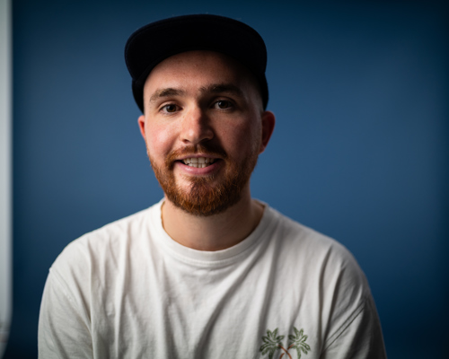 A smiling young white man with a baseball cap on looking at the camera lens